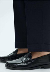 Černé kožené mokasíny s hladkým povrchem a dekorativním prošíváním, spárované s tmavě modrými kalhotami s manžetou.