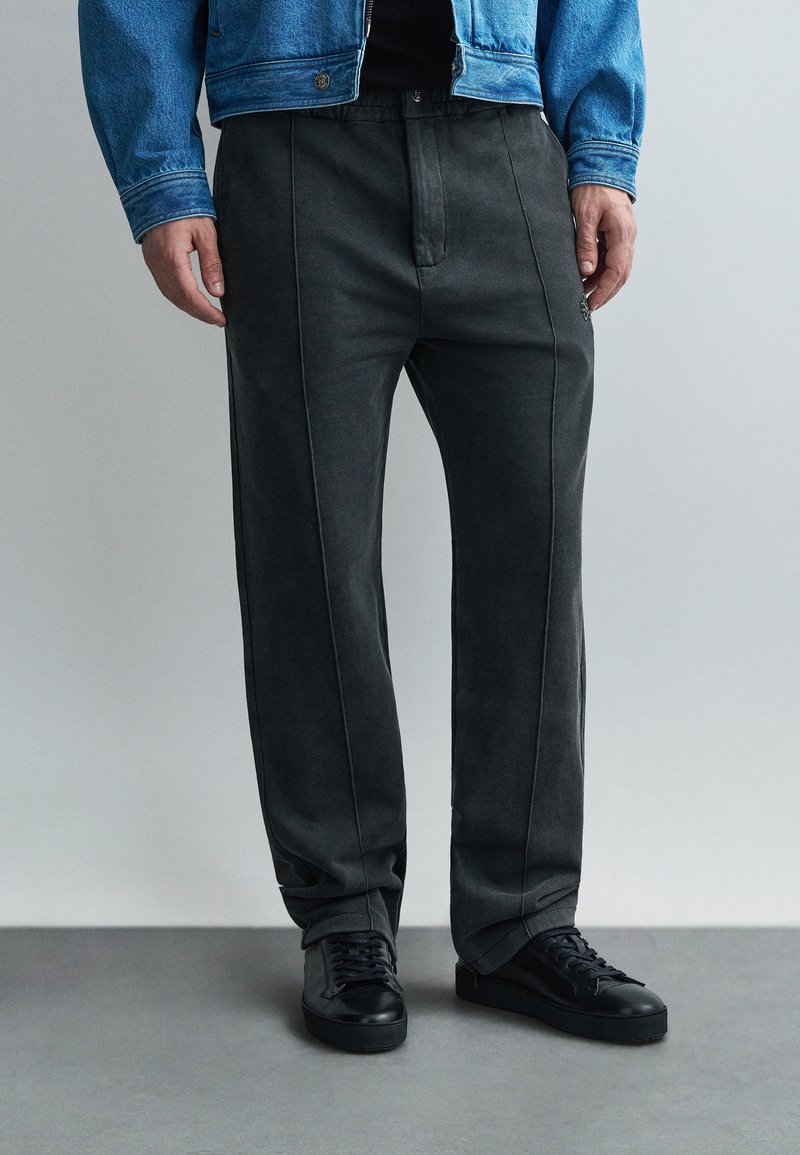 Pantalones gris oscuro hechos de tela suave, con costuras laterales, corte recto y combinados con zapatillas negras. Diseño minimalista.