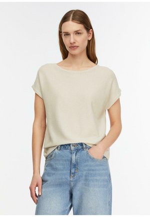 Kobieta o prostych brązowych włosach nosi beżową bluzkę z krótkim rękawem i jasnoniebieskie dżinsy z wysokim stanem, jedną ręką w kieszeni, na jednolitym białym tle.