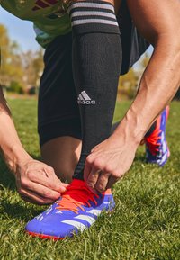 Blauwe en oranje voetbalschoenen met witte strepen, die door een hand worden vastgebonden. Zwarte sokken met strepen zichtbaar boven de enkel. Groene grasachtergrond.