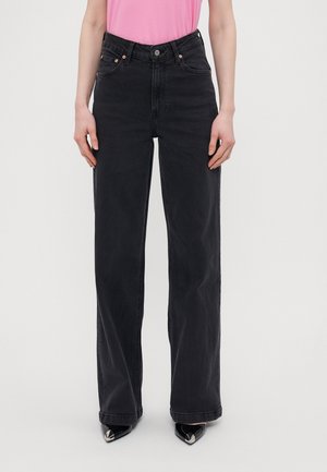 Person iført sorte jeans med høj talje og vide ben med forlommer, kombineret med en pink top og sorte spidse sko.
