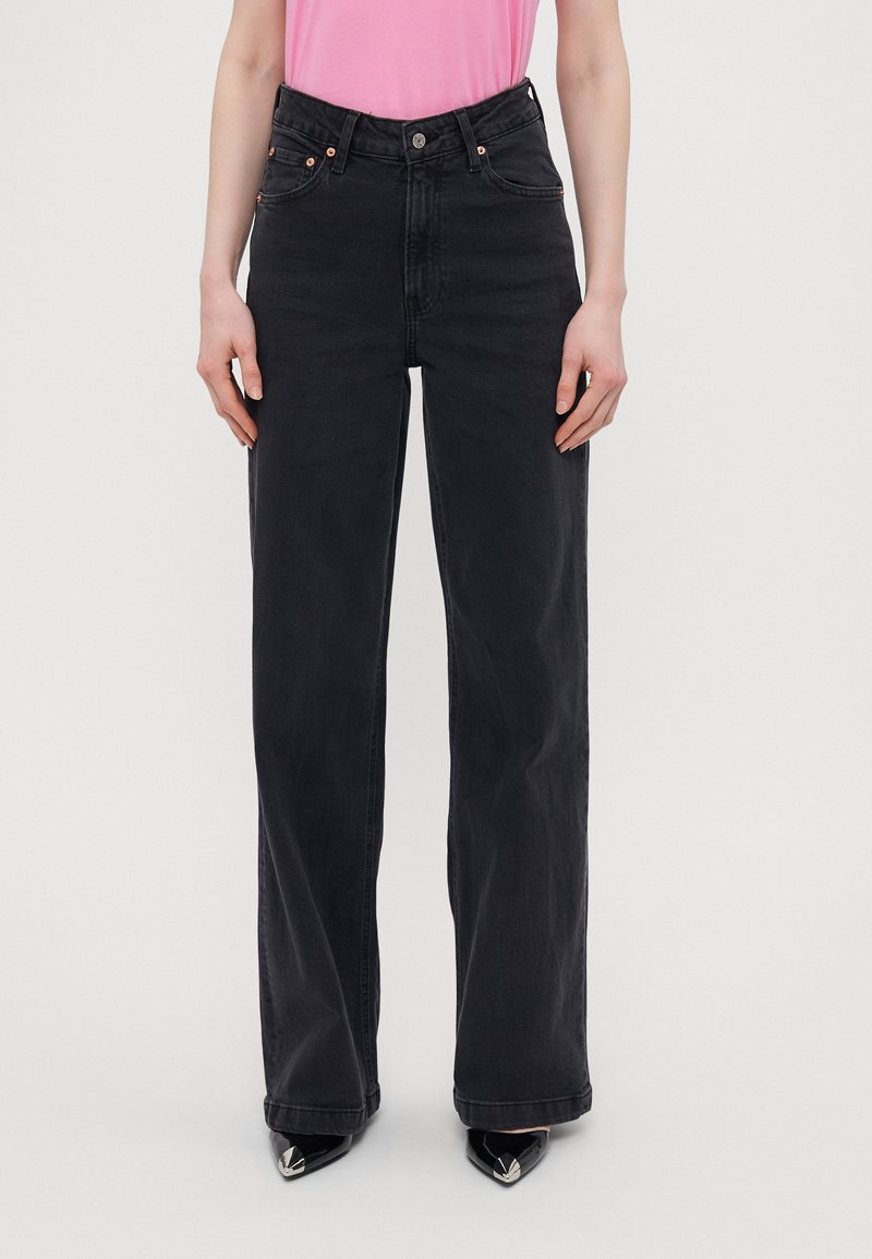 Personne portant un jean noir taille haute, jambes larges, avec des poches avant, associé à un haut rose et des chaussures noires à bout pointu.
