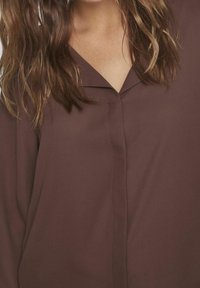 Brun blus med V-ringning, långa ärmar och en subtil glans. Den har en framfick och består av ett mjukt, lätt tyg.