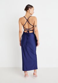 Navy blue satin maxi dress with a strappy back, featuring crisscross straps and a tied waist. Smooth texture and fitted silhouette.