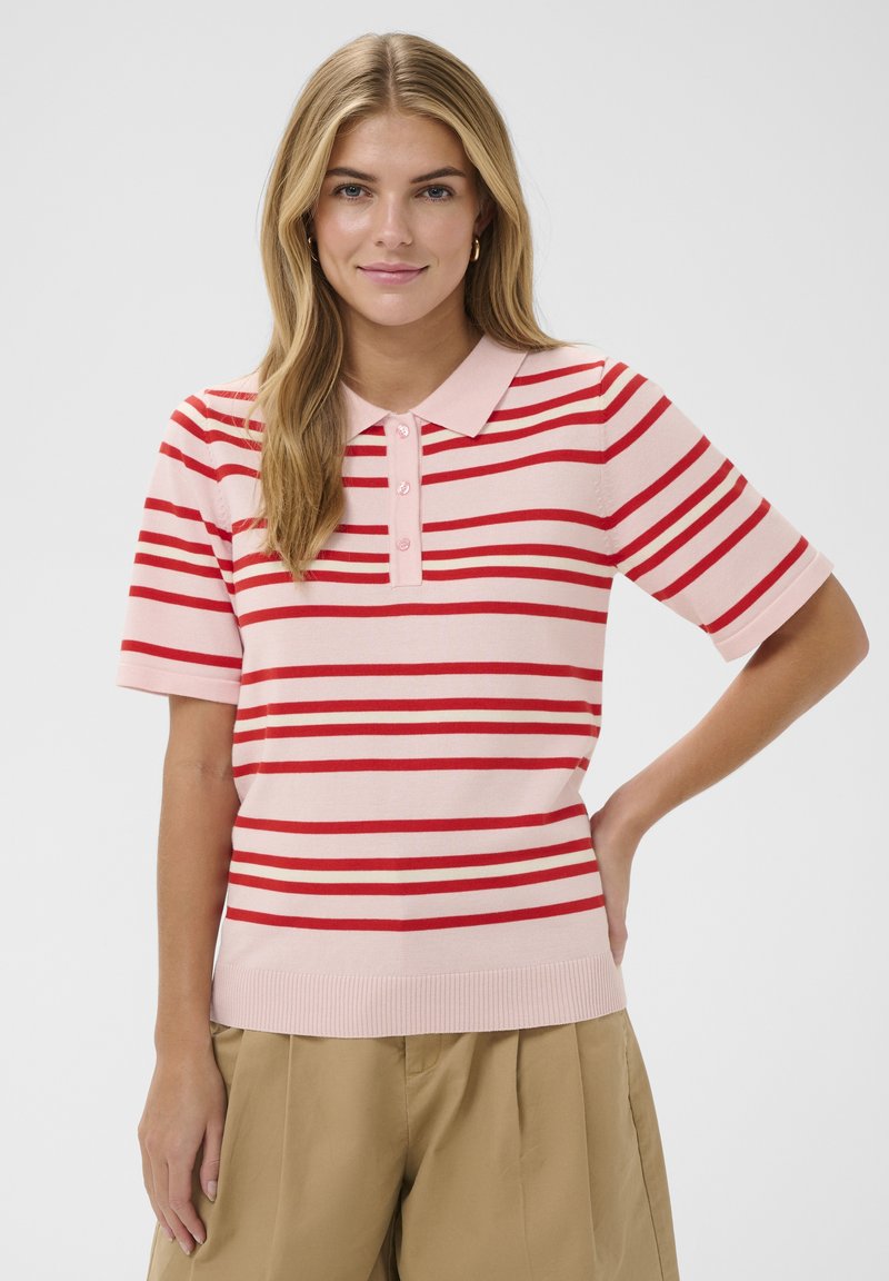 Young woman with long blonde hair wearing a pink and red striped short-sleeve polo shirt and beige pants against a plain background.