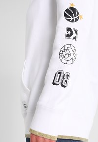 Manche de sweat-shirt blanc avec des logos de basketball noir et or et des étoiles, ainsi que le numéro 08 en lettres noires en gras près du poignet.