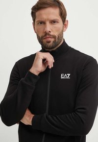 Giacca nera con zip realizzata in tessuto liscio. Presenta un colletto alto, polsini a costine e un logo EA7 bianco sul petto. Design semplice e aderente.