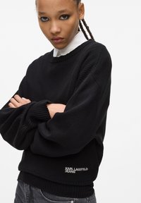 Must mustriba, millel on laiad varrukad, kaunistatud brodeeritud "Karl Lagerfeld Jeans" logoga hemmetes. Kannab valge kraega särke.