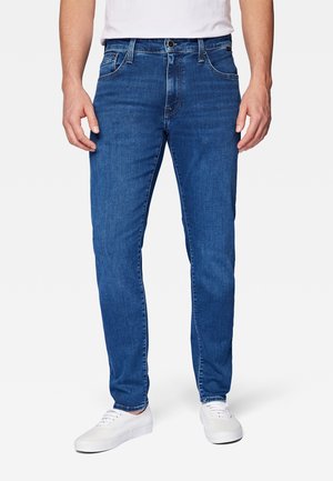 Uomo con jeans blu aderenti e sneakers bianche, in piedi su uno sfondo chiaro e semplice.