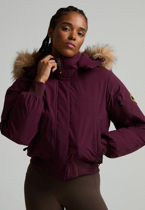 Femme aux cheveux tressés portant une veste d’hiver bordeaux à capuche avec bordure en fourrure, tenant la fermeture éclair près du cou sur un fond uni.