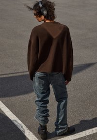 Personne avec des cheveux bouclés portant des écouteurs, un pull marron, un jean ample et des bottes noires, se tenant sur un asphalt avec des lignes de stationnement visibles.