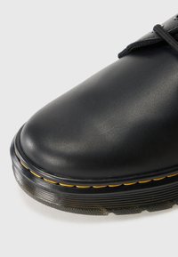 Black leather boot toe with yellow stitching and thick grooved sole on a white background.