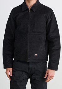 Mężczyzna w czarnej kurtce na zamek z korduroyu i czarnych spodniach cargo, stojący na tle gładkiej, jasnej ściany.