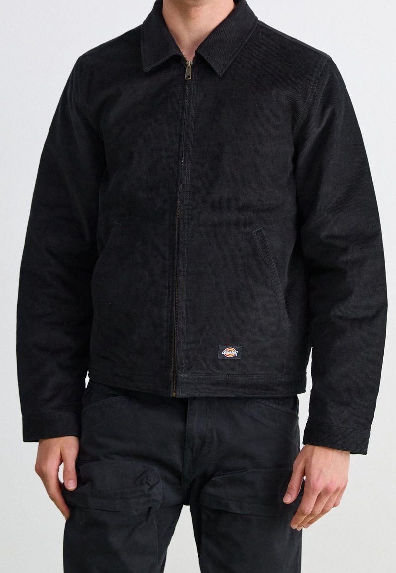 Mężczyzna w czarnej kurtce na zamek z korduroyu i czarnych spodniach cargo, stojący na tle gładkiej, jasnej ściany.