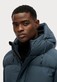 Chaqueta acolchada de color azul marino con un gran cuello, cremallera visible y capucha ajustable con cordón. Presenta un cuello alto negro texturizado debajo.