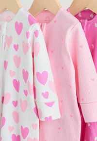 Trzy ubranka dla niemowląt: białe z dużymi różowymi sercami oraz dwa różowe z mniejszymi wzorami serc, wszystkie wykonane z prążkowanej tkaniny z długimi rękawami.