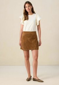 Weißes Oberteil aus Stoff mit Rüschensleeves, kombiniert mit einem braunen Wildleder-Minirock mit zwei Vordertaschen. Ballerinas im Schlangenhautmuster runden das Outfit ab.