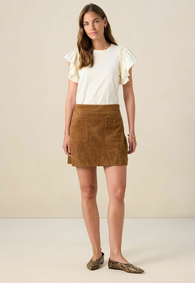 Weißes Oberteil aus Stoff mit Rüschensleeves, kombiniert mit einem braunen Wildleder-Minirock mit zwei Vordertaschen. Ballerinas im Schlangenhautmuster runden das Outfit ab.