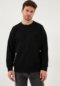 Črna sweatshirt iz mehkega materiala, s krožnim izrezom, raglan rokavi in majhnim, okroglim logotipom na prsih.