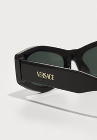 Gafas de sol rectangulares negras con lentes oscuros y montura gruesa. "VERSACE" está grabado en dorado en el exterior de la patilla.