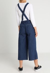 Denimoverall med vid bendesign, mörkblå färg, korsade bärremmar och två bakfickor. Matchad med svarta sneakers.