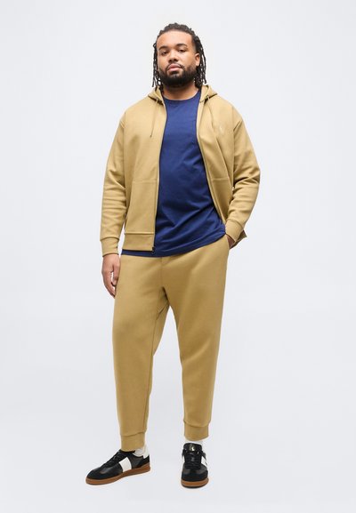 Άνδρας με dreadlocks που φοράει μπεζ φούτερ με κουκούλα και παντελόνι φόρμας, μπλε σκούρο μπλουζάκι, μαύρα και άσπρα αθλητικά παπούτσια, στέκεται μπροστά σε λευκό φόντο.