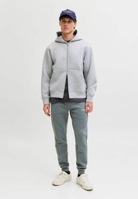 Graue Zip-Hoodie mit Kängurutaschen, kombiniert mit hellgrauen Trainingshosen mit Seitenpaneelen. Das Model trägt eine marineblaue Mütze und weiße Sneaker.