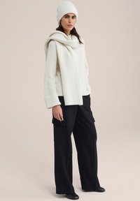 Maglione bianco a coste con collo drappeggiato, abbinato a pantaloni cargo neri a gamba larga con tasche laterali e un berretto coordinato.