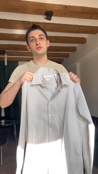 Νέος άνδρας που κρατάει ένα γκρι και άσπρο ριγωτό πουκάμισο με κουμπιά, στέκεται σε εσωτερικό χώρο με ξύλινες δοκούς οροφής και σύγχρονο φωτισμό από πάνω.