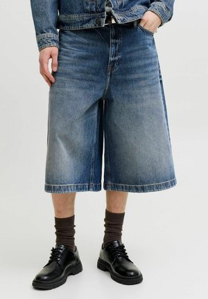 Homme portant un short en jean bleu large arrivant aux genoux, des chaussettes foncées, des chaussures en cuir noires et une veste en jean bleu avec une main dans la poche.