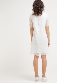 Valkoinen pitsimekko, jossa lyhyet hihat, kukkakuvio, aaltoileva helma ja vetoketju selässä. Yhdistetty valkoisiin liukuvaan malliin tehtyihin lenkkareihin.