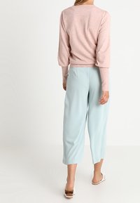 Žena oblečená v světle růžovém svetru s dlouhým rukávem a volných světle modrých 3/4 kalhotách, stojí s obratem zády k divákovi.