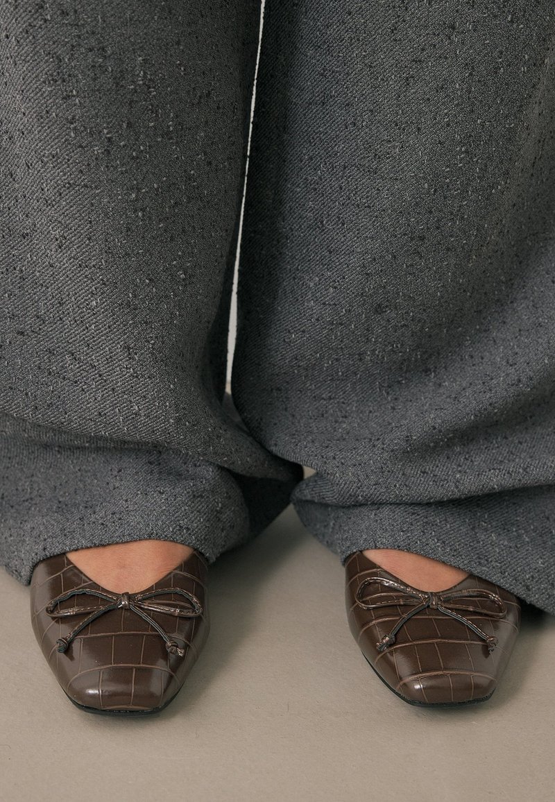 Ballerines marron texturées avec des nœuds, portées avec un pantalon gris ample couvrant les chevilles, debout sur un sol lisse.