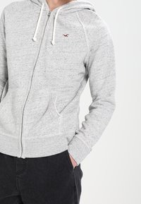 Person som bär en ljusgrå zip-up huvtröja med framfickor och en liten röd logotyp på vänster bröst, i kombination med svarta byxor.