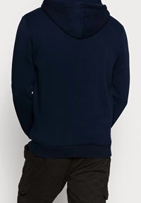 Sudadera con capucha de color azul marino, con una textura suave, puños y dobladillo acanalados, que cuenta con un bolsillo tipo canguro. La vista trasera muestra la capucha y las costuras.