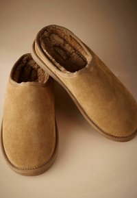 Brown suede slippers with a soft, fuzzy interior lining and a slip-on design, featuring stitched accents and a flat sole.