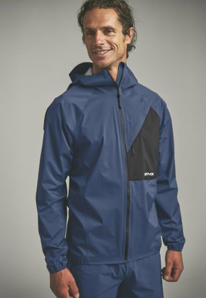 Mann trägt eine blaue Regenjacke mit schwarzer Brusttasche, Kapuze und durchgehendem Reißverschluss, lächelt und schaut leicht nach rechts.