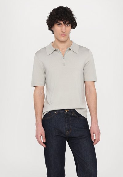Polo grigio chiaro a maniche corte con colletto con zip, abbinato a jeans blu scuro. Il tessuto appare liscio con una lieve trama.