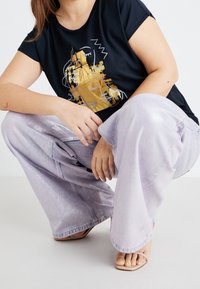 Black T-shirt with a gold and white graphic design. Light purple wide-leg pants with a shiny texture. Neutral sandals visible.