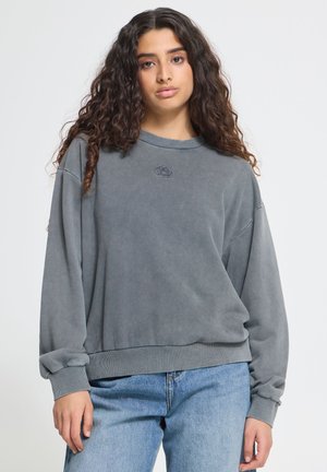 Kvinde med langt krøllet hår iført en løs grå sweatshirt og blå jeans, stående mod en ensfarvet lys baggrund.