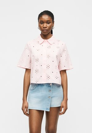 Nő világos rózsaszín lyukacsos rövid ujjú ingben és világoskék aszimmetrikus, gombos farmer szoknyában áll egy sima háttér előtt.