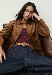 Žena s dlhými tmavými vlasmi opretá o hnedé kožené kreslo, oblečená v hnedej semišovej bunde, bordovom tričku, širokých tmavých džínsach a hnedom opasku so zlatou sponou.