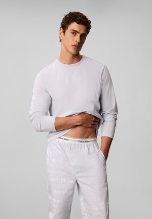 Homme aux cheveux bouclés portant une chemise légère à manches longues et un pantalon à motifs soulève sa chemise pour montrer la ceinture de son sous-vêtement de marque Calvin Klein.
