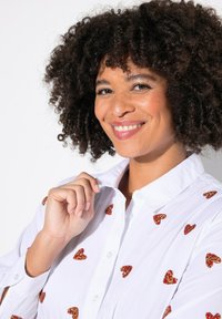 Camicia bianca con bottoni e paillettes a forma di cuore rosse e dorate distribuite uniformemente sulla stoffa. Sono visibili il colletto e le maniche lunghe.