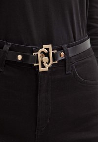 Cintura in pelle nera con fibbia in metallo dorato caratterizzata da un design intrecciato. Design semplice con piccoli accenti hardware dorati.