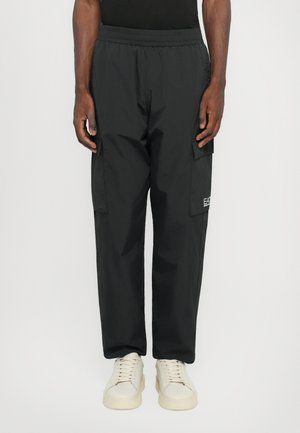 Pantalons cargo noirs avec une taille élastique, des poches latérales et un tissu texturé. Présente un logo sur la jambe droite. Portés avec des baskets claires.