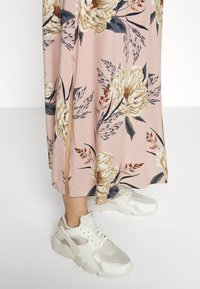 Abito lungo floreale con sfondo rosa, caratterizzato da fiori bianchi e blu, abbinato a sneaker bianche chunky. L'abito ha uno spacco laterale.