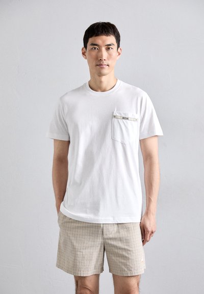 Uomo che indossa una camicia bianca a maniche corte con taschino e pantaloni corti a quadri beige, in piedi davanti a uno sfondo grigio chiaro uniforme.