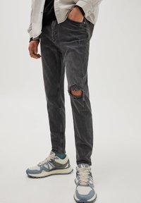 Vaqueros negros rasgados con un corte ajustado. Presenta bordes deshilachados y acentos desgastados. Combinados con zapatillas de color claro y una blusa con capas.
