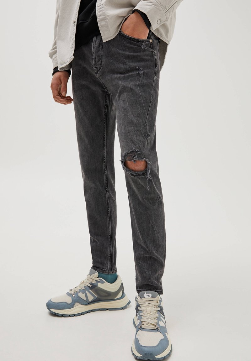 Vaqueros negros rasgados con un corte ajustado. Presenta bordes deshilachados y acentos desgastados. Combinados con zapatillas de color claro y una blusa con capas.